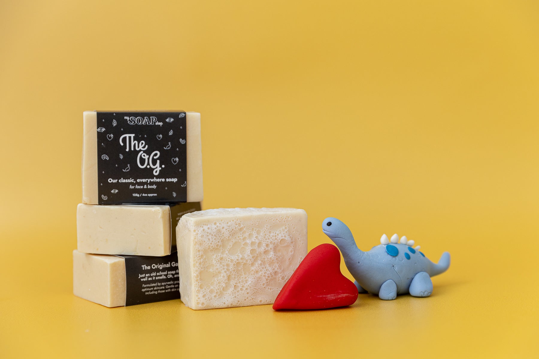 four bars of The OG soap with toy dinosaur and red love heart on yellow background